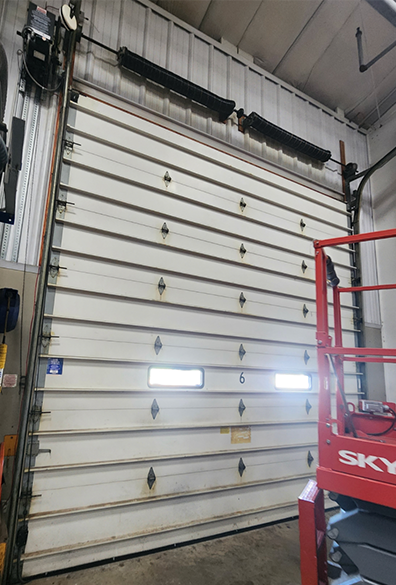 Closed white garage door inside an industrial warehouse, with a red scissor lift beside it.