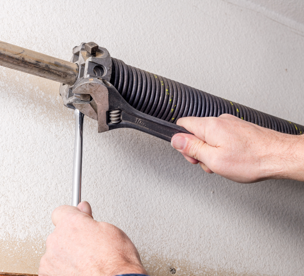 Hands using a wrench to adjust a garage door spring mechanism on a wall-mounted track.