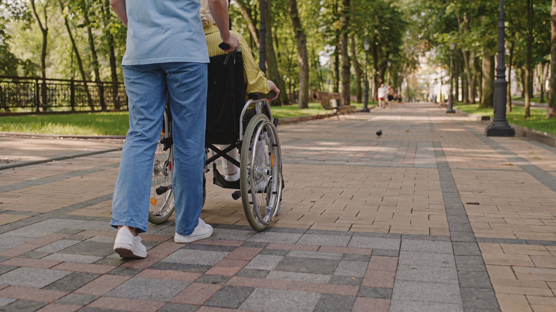 An individual in a wheelchair being pushed down a walkway by someone in jeans