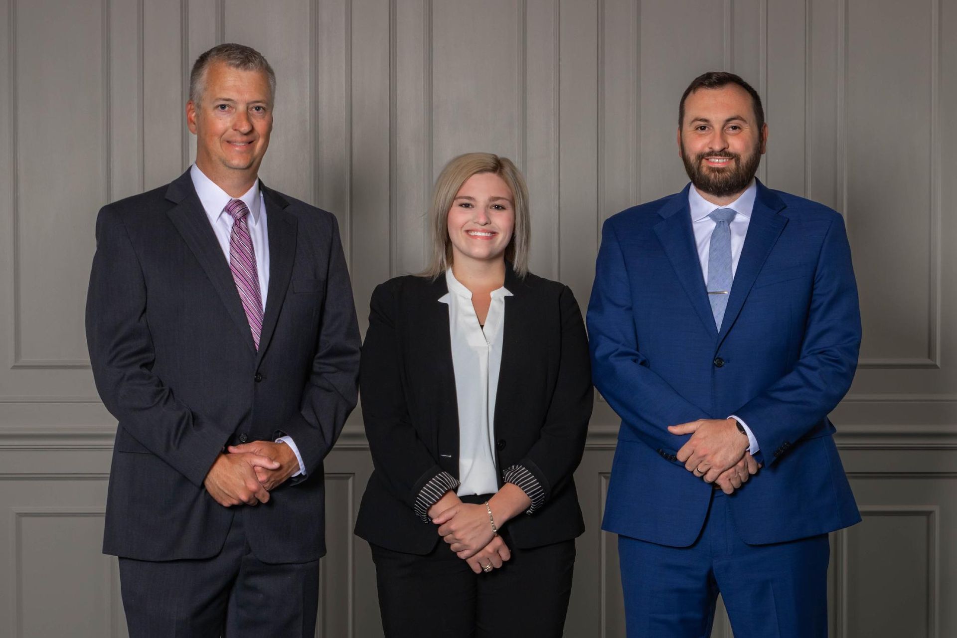 Three professionals in business attire stand side-by-side against a gray paneled wall, smiling at the camera.