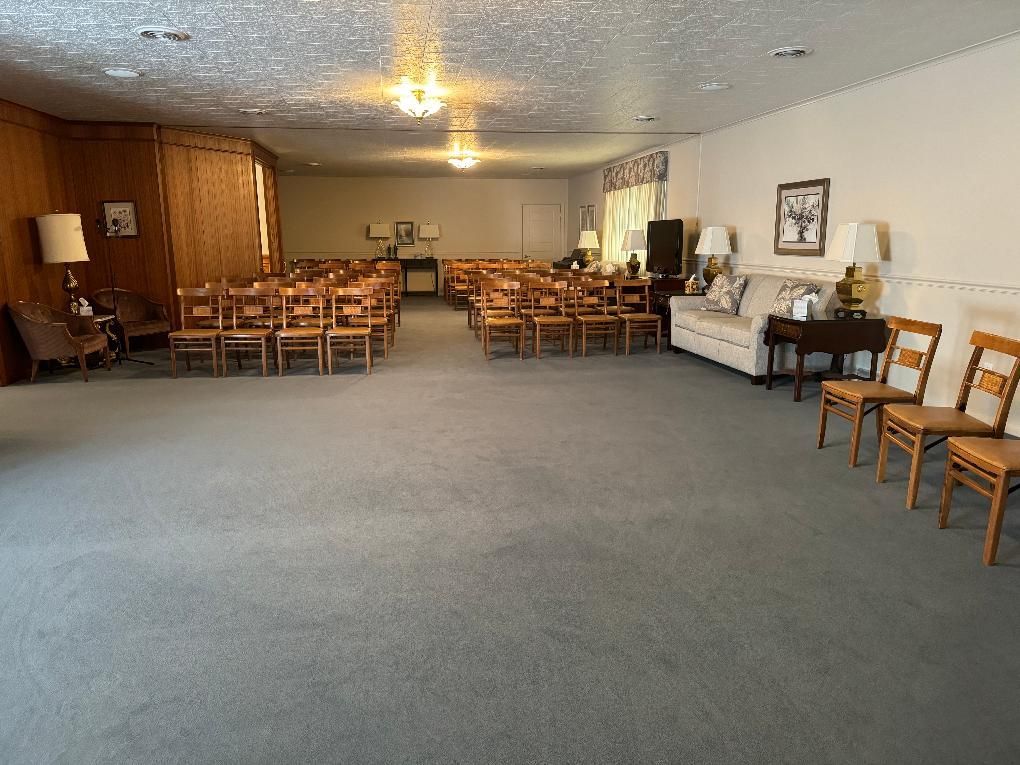 An indoor event space with wooden chairs arranged in rows facing the back, a light-colored sofa, and grey carpeting.