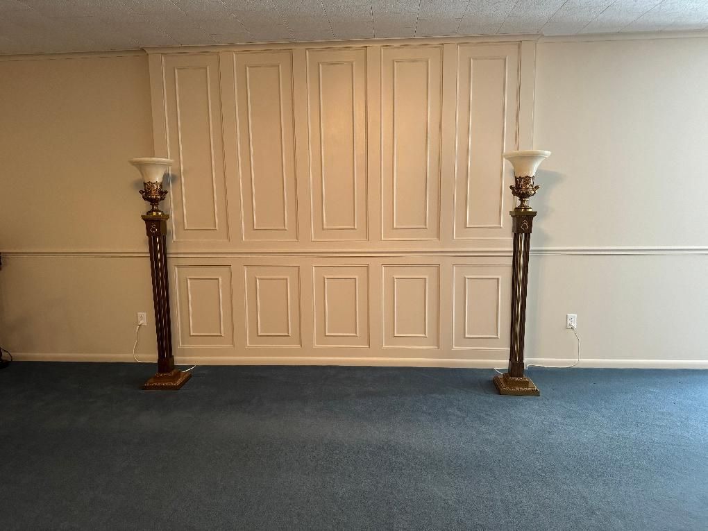 A cream-colored paneled wall features two ornate bronze floor lamps on blue carpet in a minimalist room.