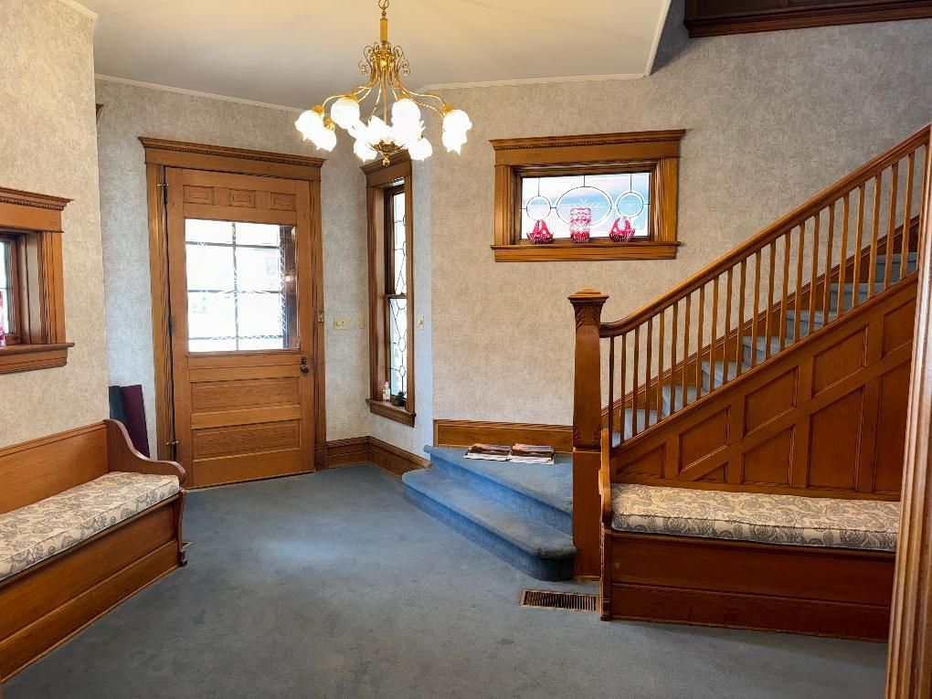 An entryway with blue carpet, wood-paneled stairs with a built-in bench, a wooden front door, and a hanging chandelier.