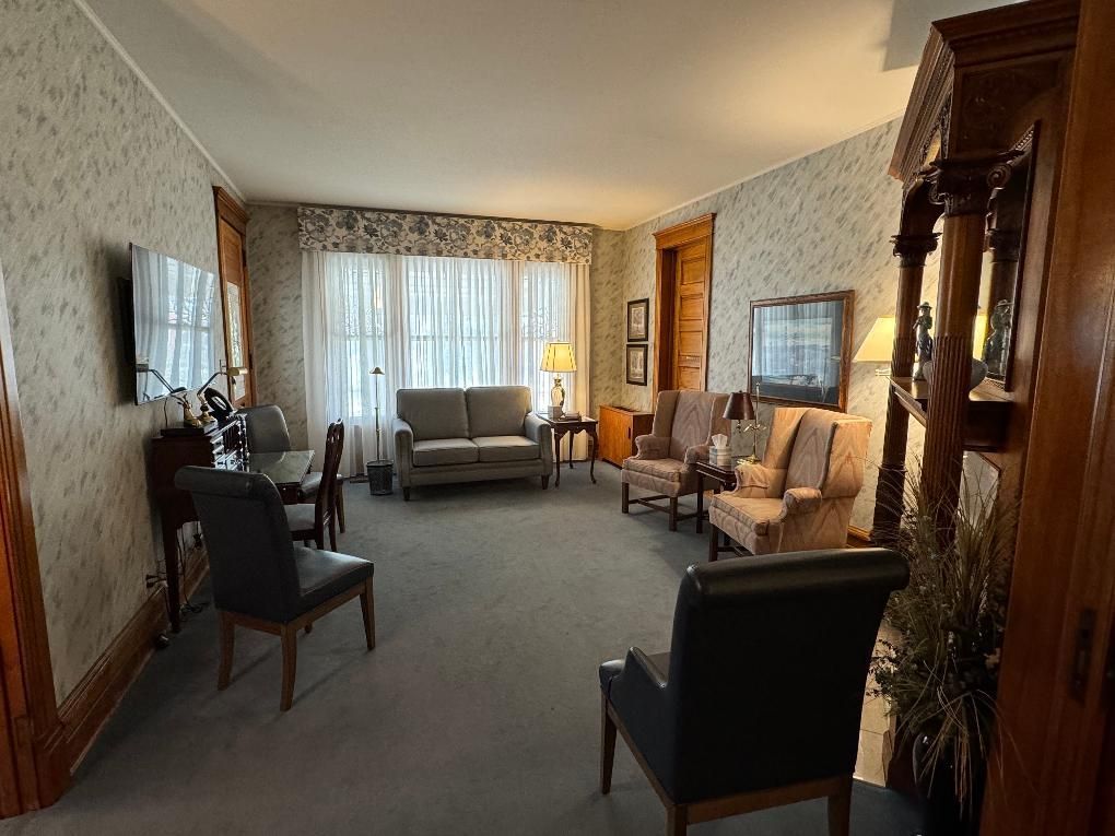 A cozy parlor with vintage wallpaper, a central sofa, upholstered chairs, a small table, and wood trim throughout.