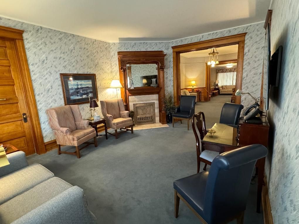 A sitting room with floral wallpaper, a fireplace, two beige armchairs, and several dark chairs arranged on gray carpet.