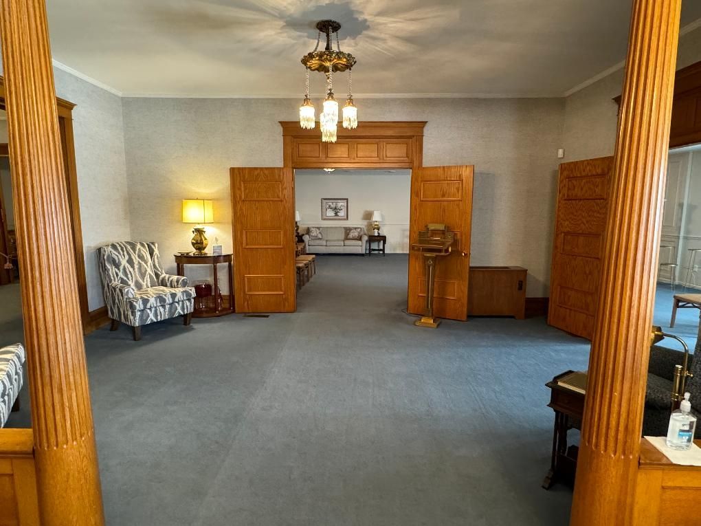 A quiet, carpeted funeral home parlor with wood-trimmed doorways, a patterned armchair, and a classic chandelier.