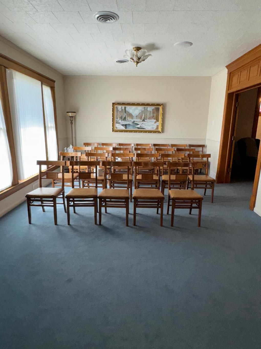 A room with rows of wooden chairs arranged in front of a landscape painting on a cream wall, with blue carpeting.