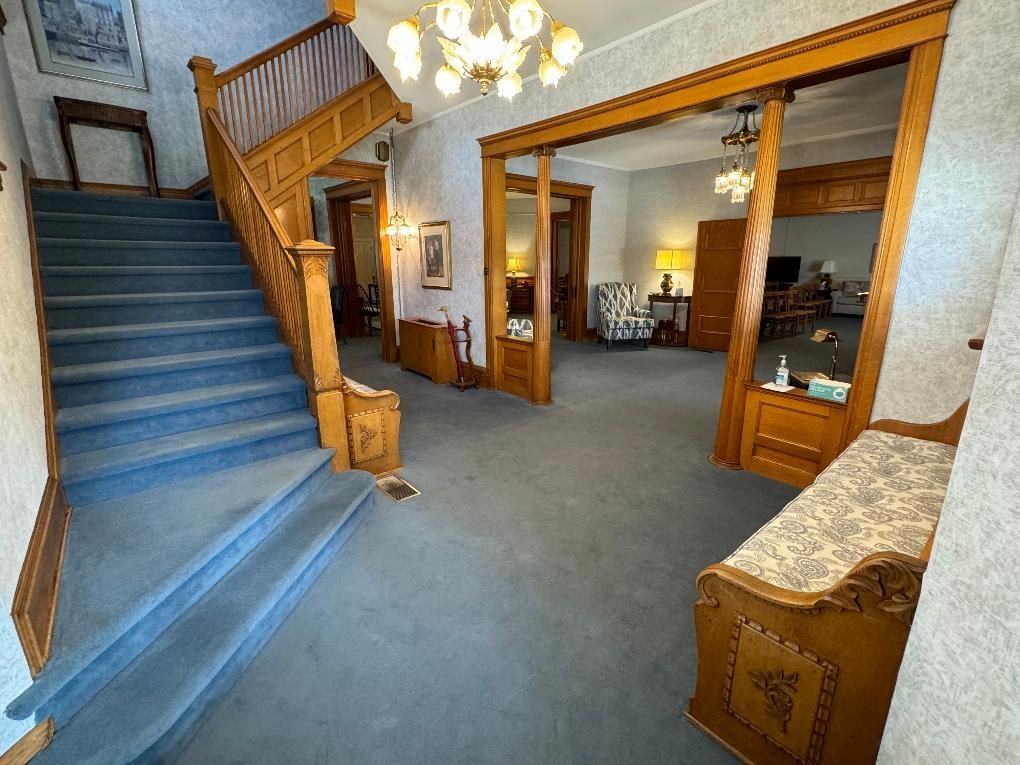 A carpeted entryway featuring a wooden staircase, a view into a living area, and a patterned bench in the foreground.