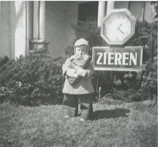 A child in a winter coat and hat holds a large object while standing on a lawn in front of a clock sign reading "ZIEREN".