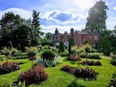 A red brick manor house stands behind a vibrant garden with manicured shrubs and flowerbeds under a bright, cloudy sky.