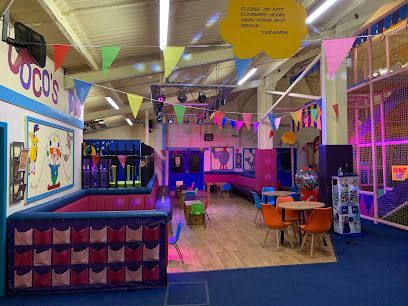 Indoor children's play area with purple-cushioned benches, wooden tables with chairs, and colorful bunting decorations.