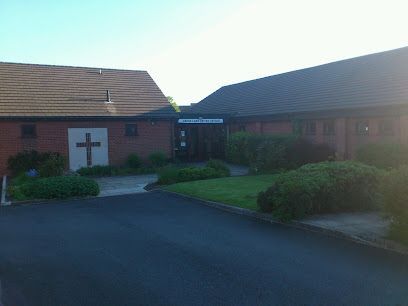 A brick church building with a cross on the left wall, a central entryway, and a patch of lawn in front.