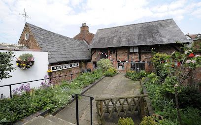 A garden path leads to an old brick building with a slate roof and decorative bunting, surrounded by lush green foliage.