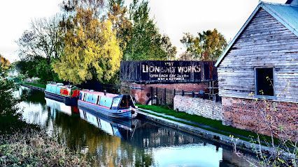 Narrowboats moored on a calm canal next to the brick and wood-sided Lion Salt Works building under autumn trees.