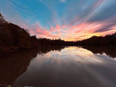 A serene lake reflects a vibrant, colorful sunset with pink and orange clouds, framed by dark silhouettes of trees.
