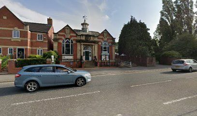 A street view of a small brick building with arched windows and a cupola, flanked by houses with cars parked out front.