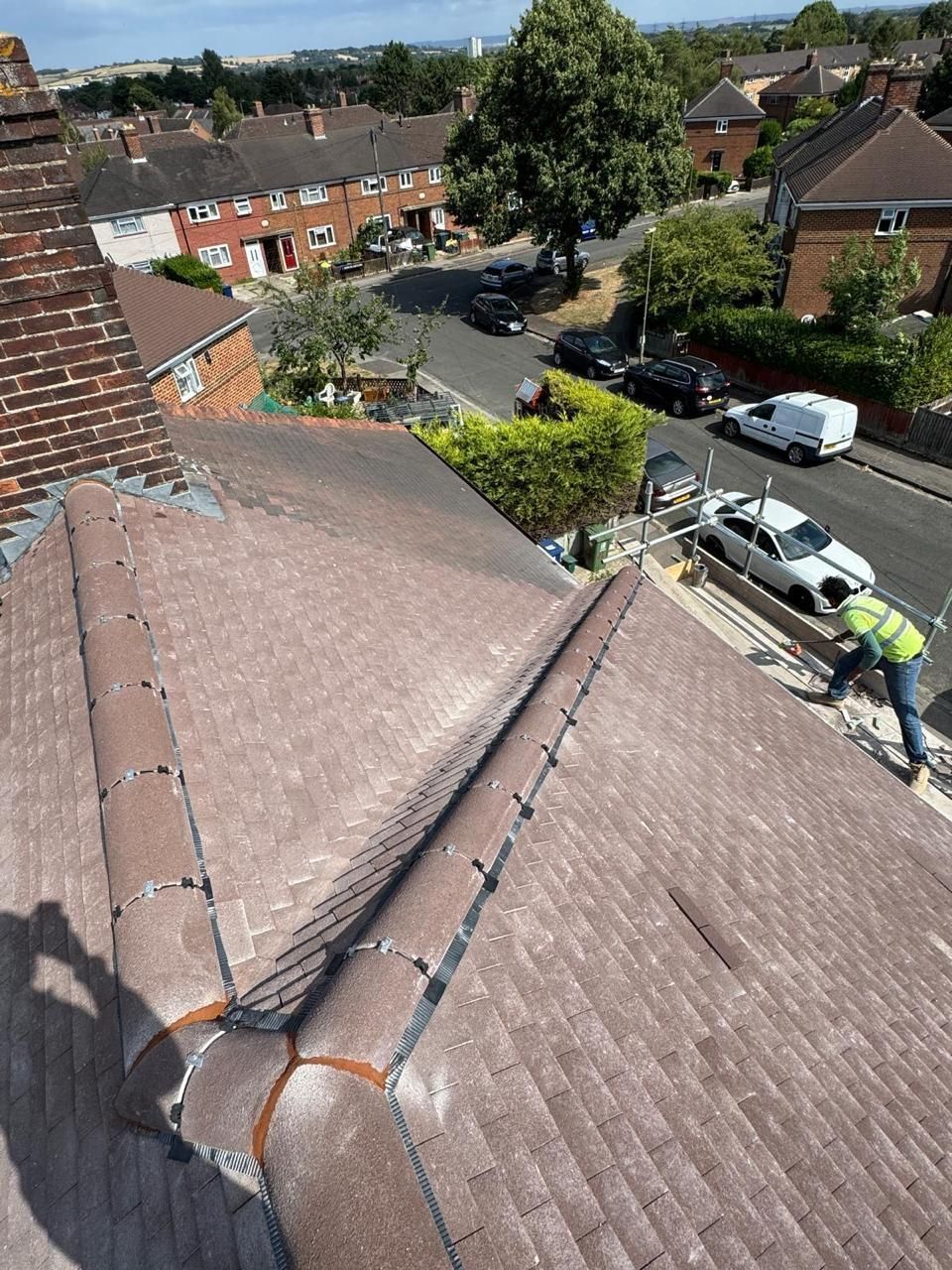 A view from a rooftop showing a worker in a high-visibility vest repairing ridge tiles on a suburban house.