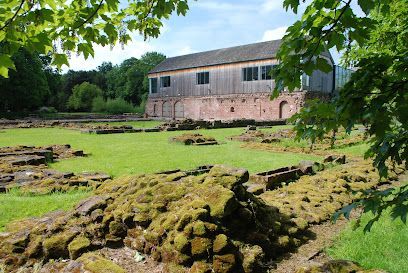 The moss-covered stone ruins of a historic site sit on a green lawn in front of a rustic wooden building with a tiled roof.