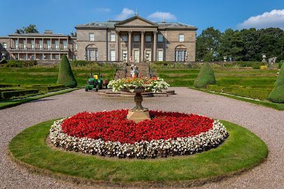 A stately stone mansion overlooks a formal garden with a large central bed of red and white flowers and a stone planter.