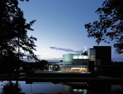 A modern building with illuminated windows reflects in dark water at twilight, framed by silhouetted trees.