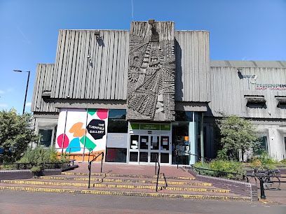 A Brutalist-style building with a central concrete relief sculpture above a glass entrance and stairs on a sunny day.