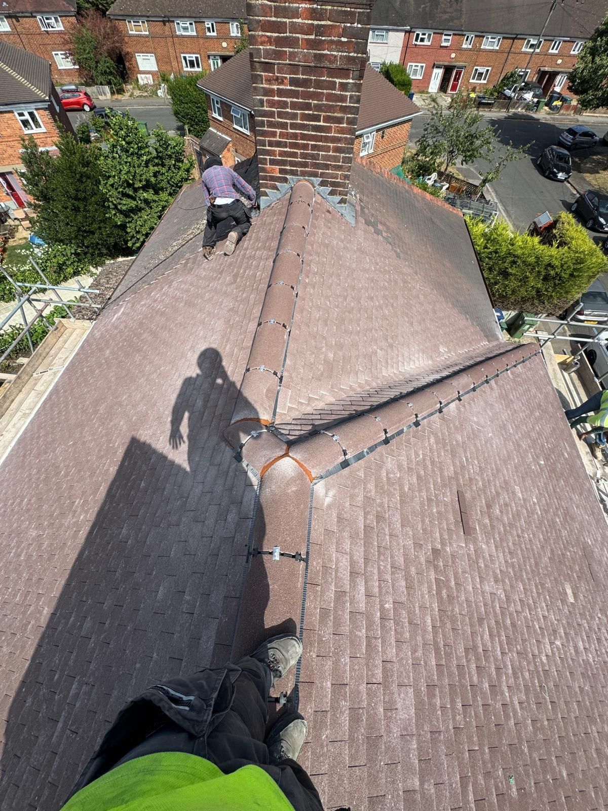 A high-angle view of two people working on a brown, shingled residential roof near a brick chimney.