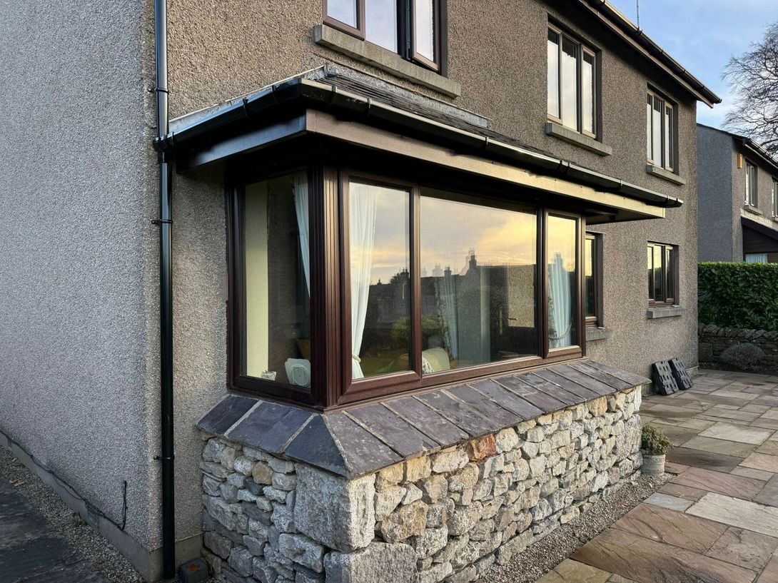 A stone-based bay window on the side of a grey pebble-dash house with a dark brown frame and a slate-tiled sill.