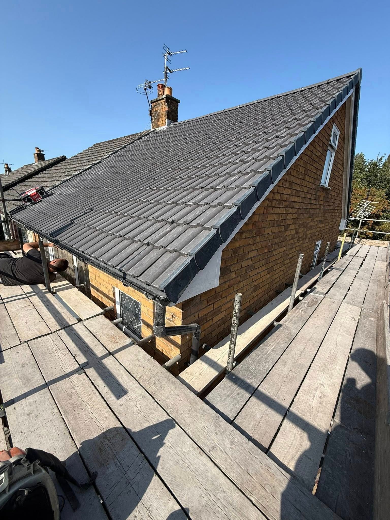 A brick house exterior under a dark tiled roof, viewed from adjacent scaffolding against a clear blue sky.