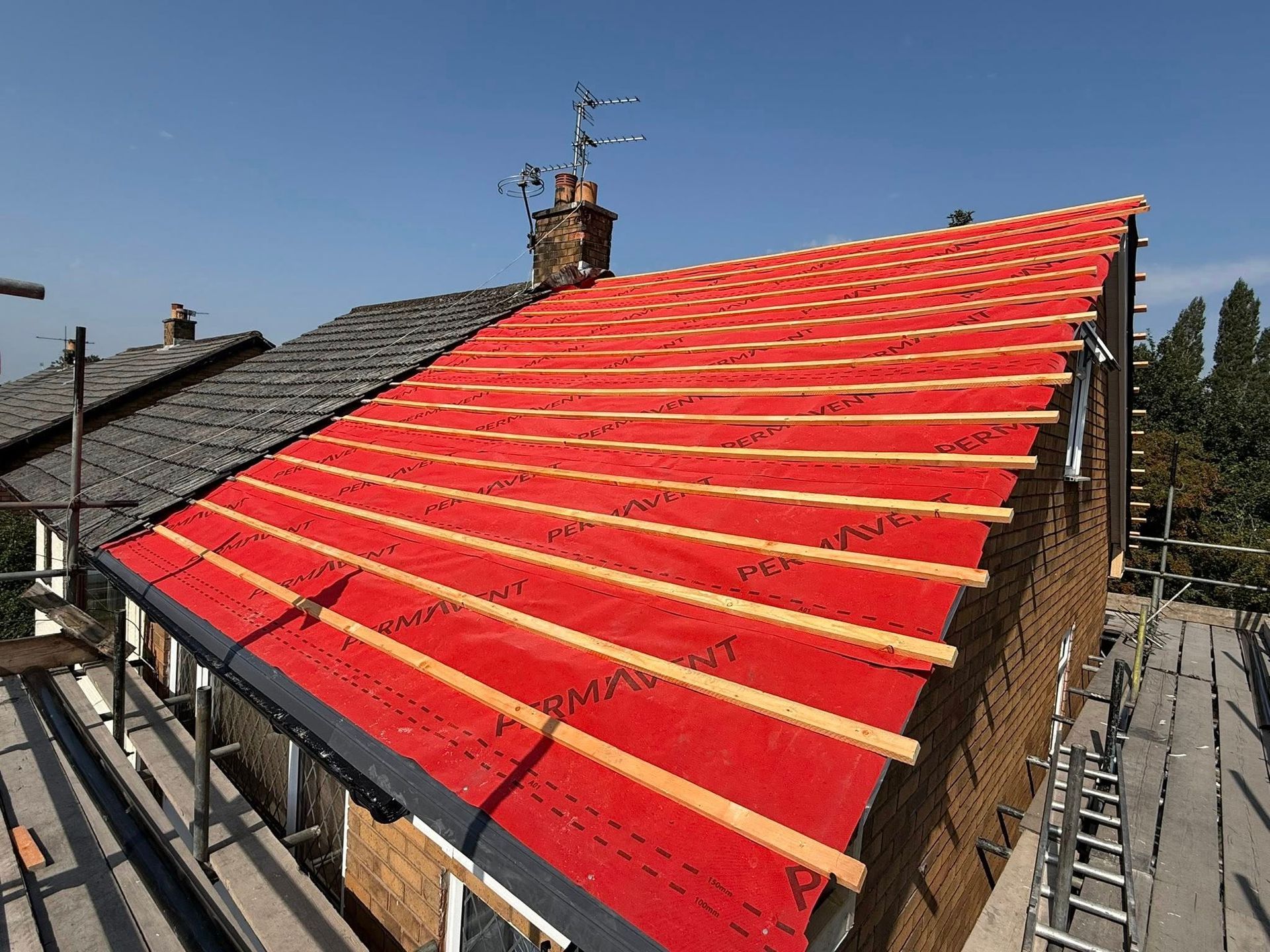 A partially renovated roof with bright red underlay and wooden battens installed, next to an old tiled roof.
