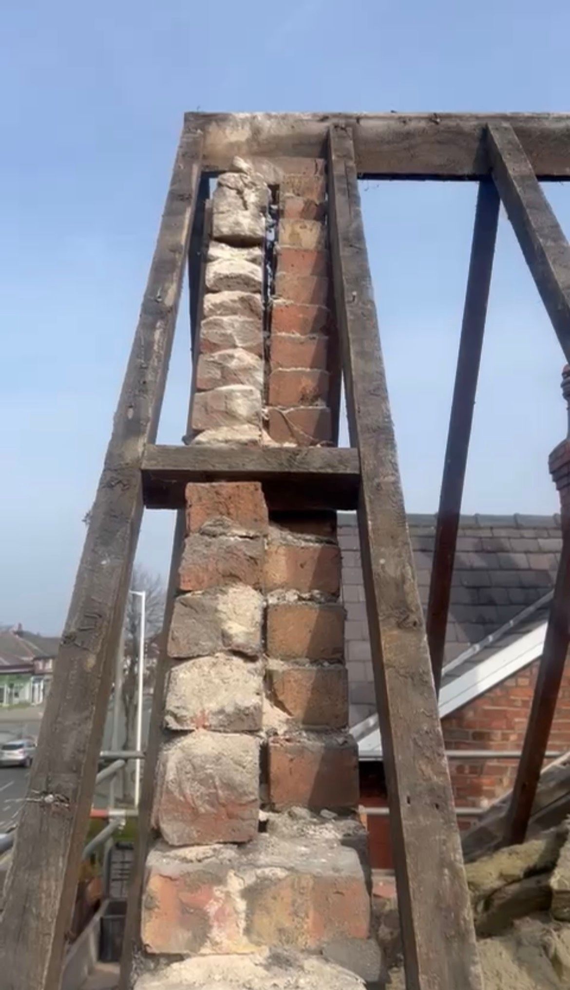 A close-up view of an unfinished brick chimney structure framed by wooden roof trusses against a clear blue sky.