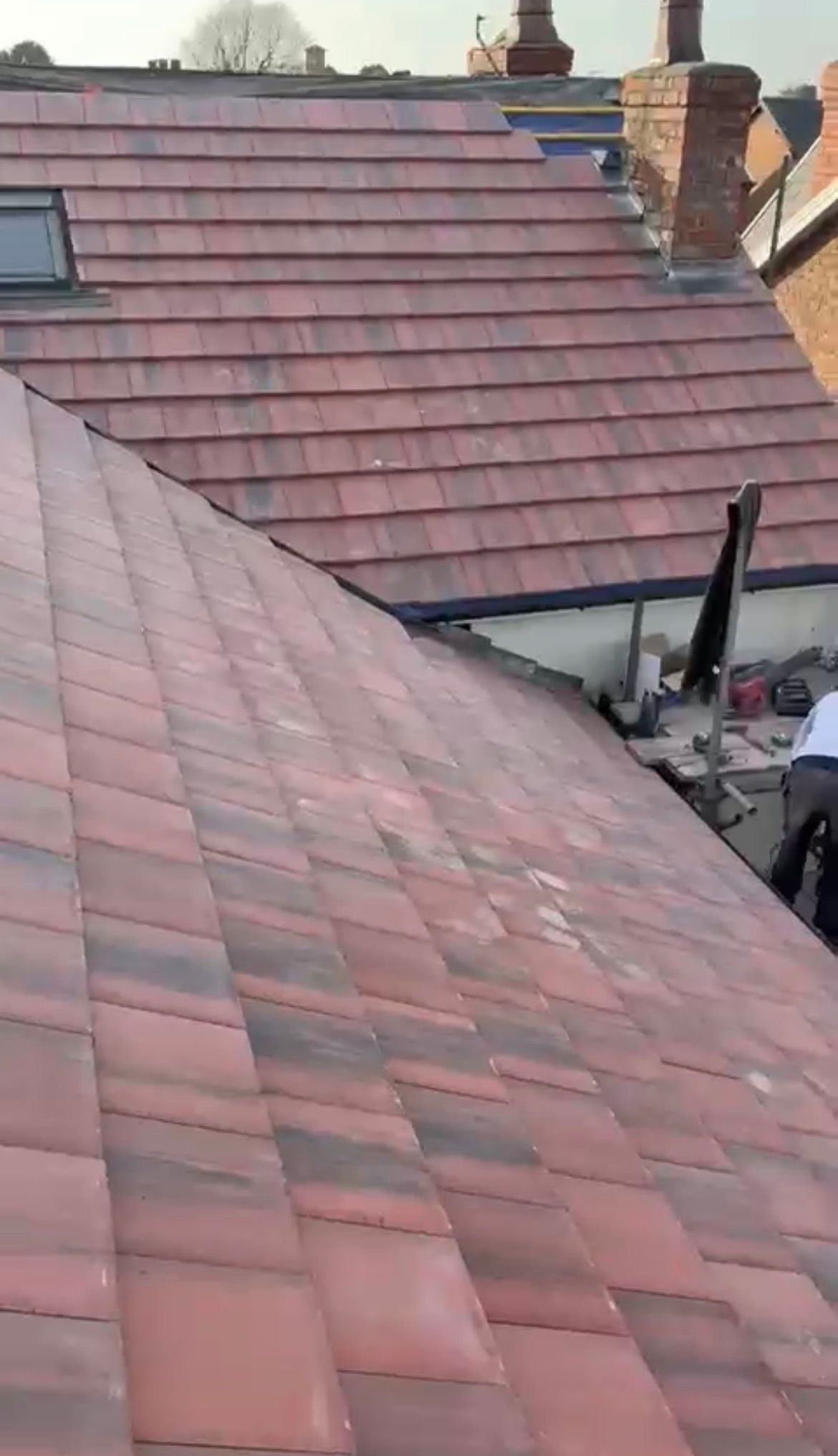 A worker on a roof in the foreground, with an adjacent tiled roof and a chimney in the background.