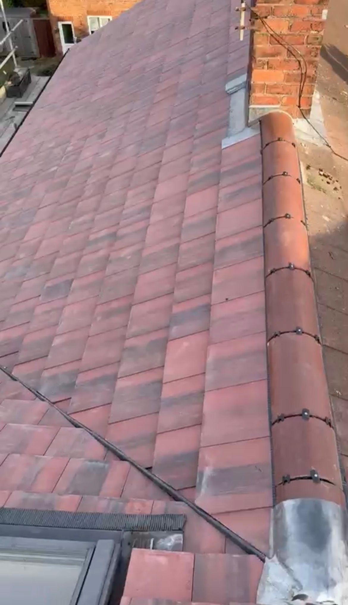 A sloping residential roof covered in red clay tiles, featuring a ridge line, a small chimney, and a skylight window.
