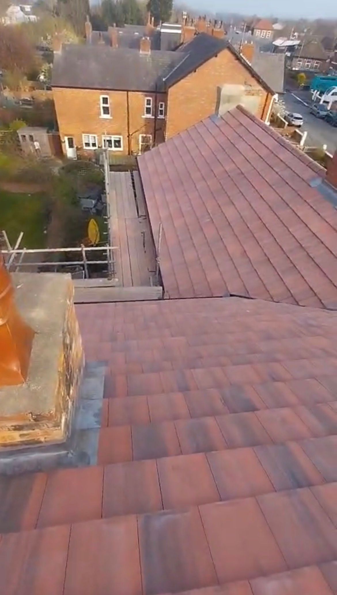 A high-angle view looking down at a newly tiled reddish-brown roof with a chimney, next to a scaffolded section.