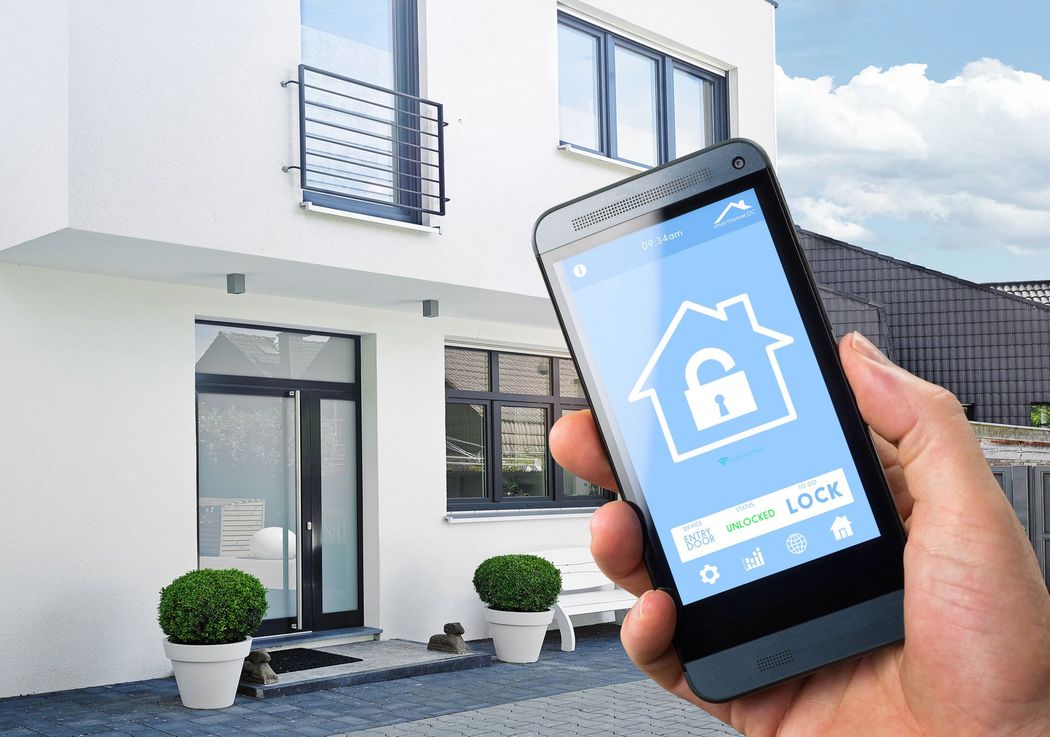 Una mano che regge uno smartphone con un'app per la sicurezza domestica davanti a una moderna casa bianca.