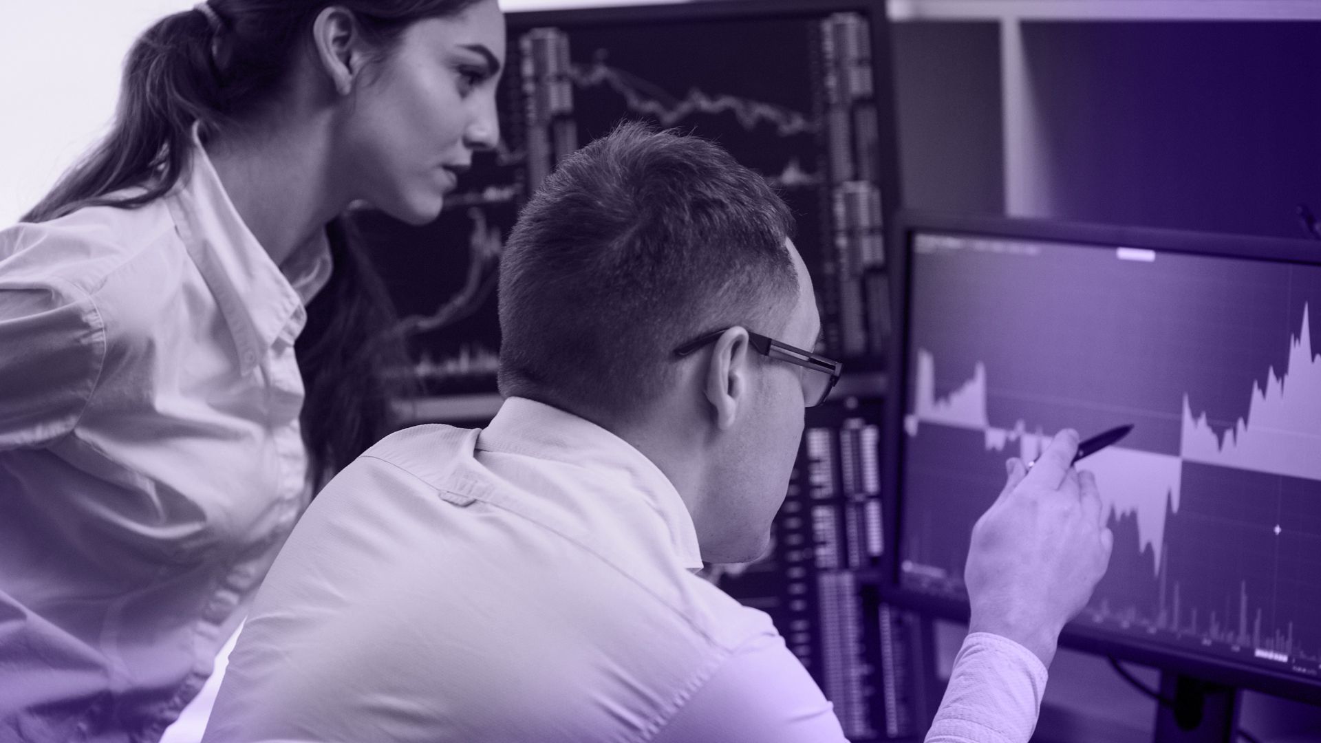 Two people looking at computer screens displaying stock charts, in a dark setting, one points.