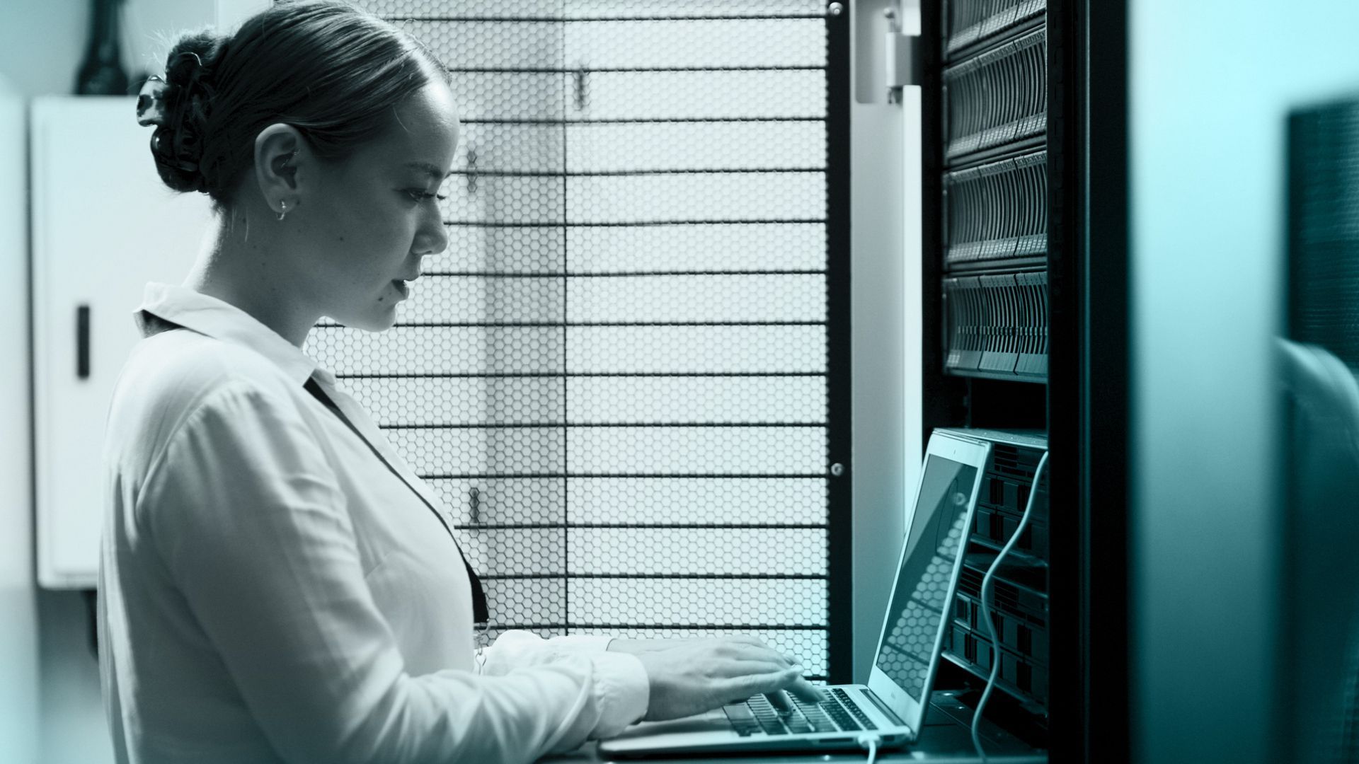 Woman working on a laptop in a server room, typing and looking at the screen.