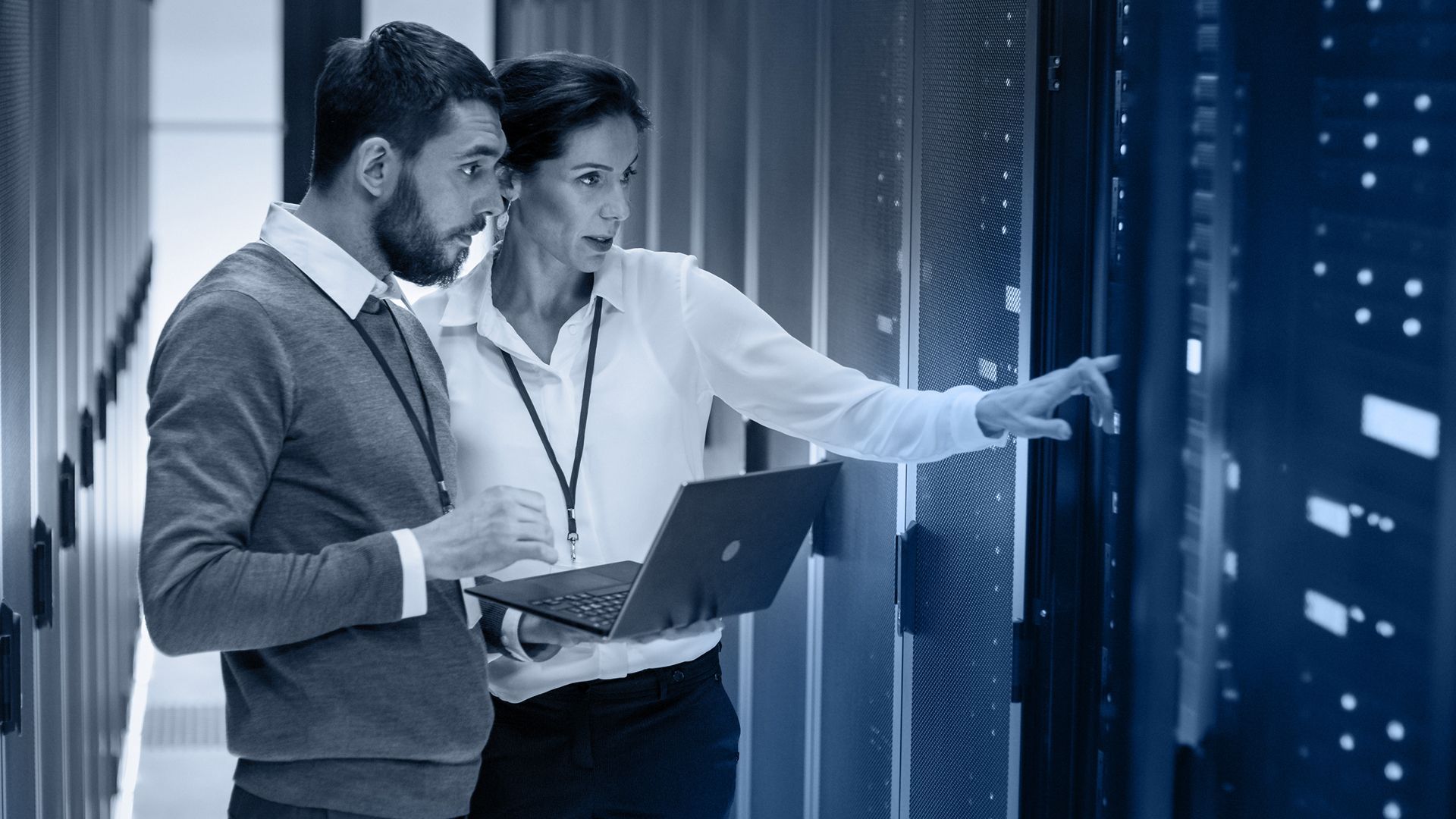 Two people in server room examining data on a panel, one pointing, holding a laptop.