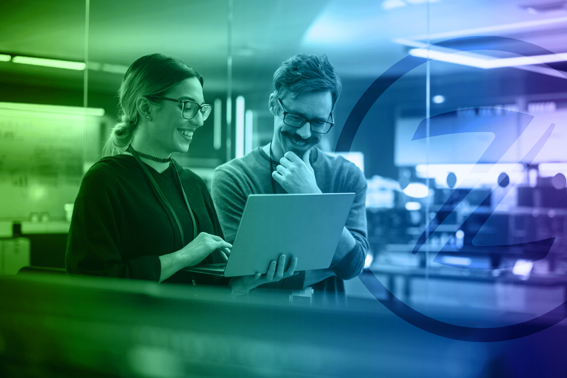 Two people looking at a laptop, smiling. Inside a modern office with green and blue lighting.