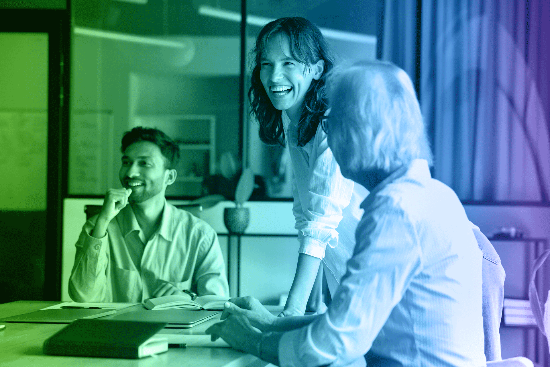 Three people laughing in an office setting around a table. One leans over, smiling. Others are seated.