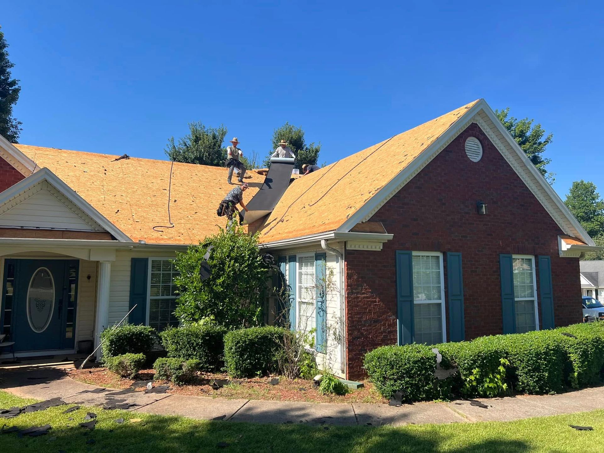 Roofers on a brick house removing old shingles under a blue sky.