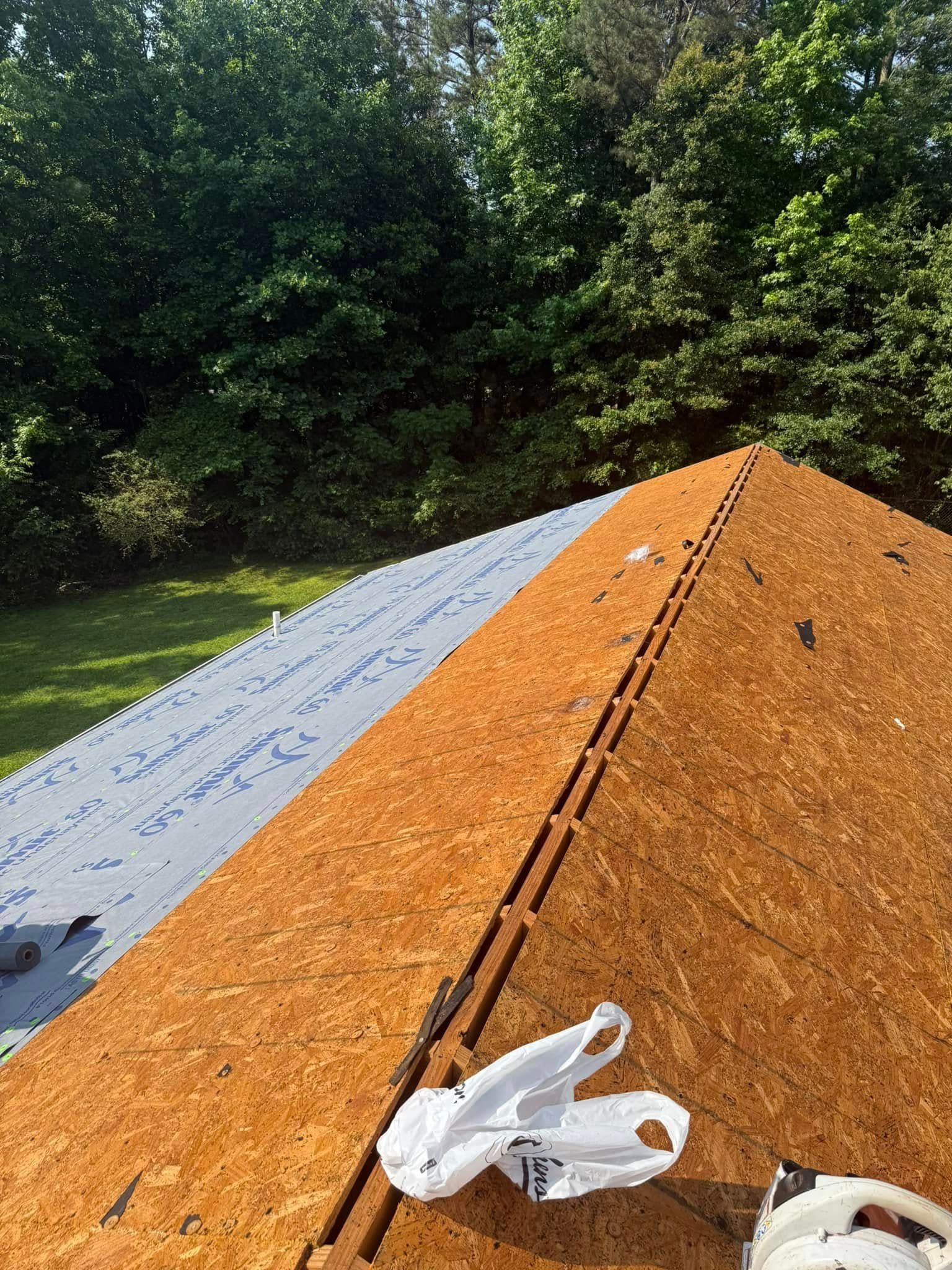 Roof partially covered with plywood and underlayment, with trees in the background.