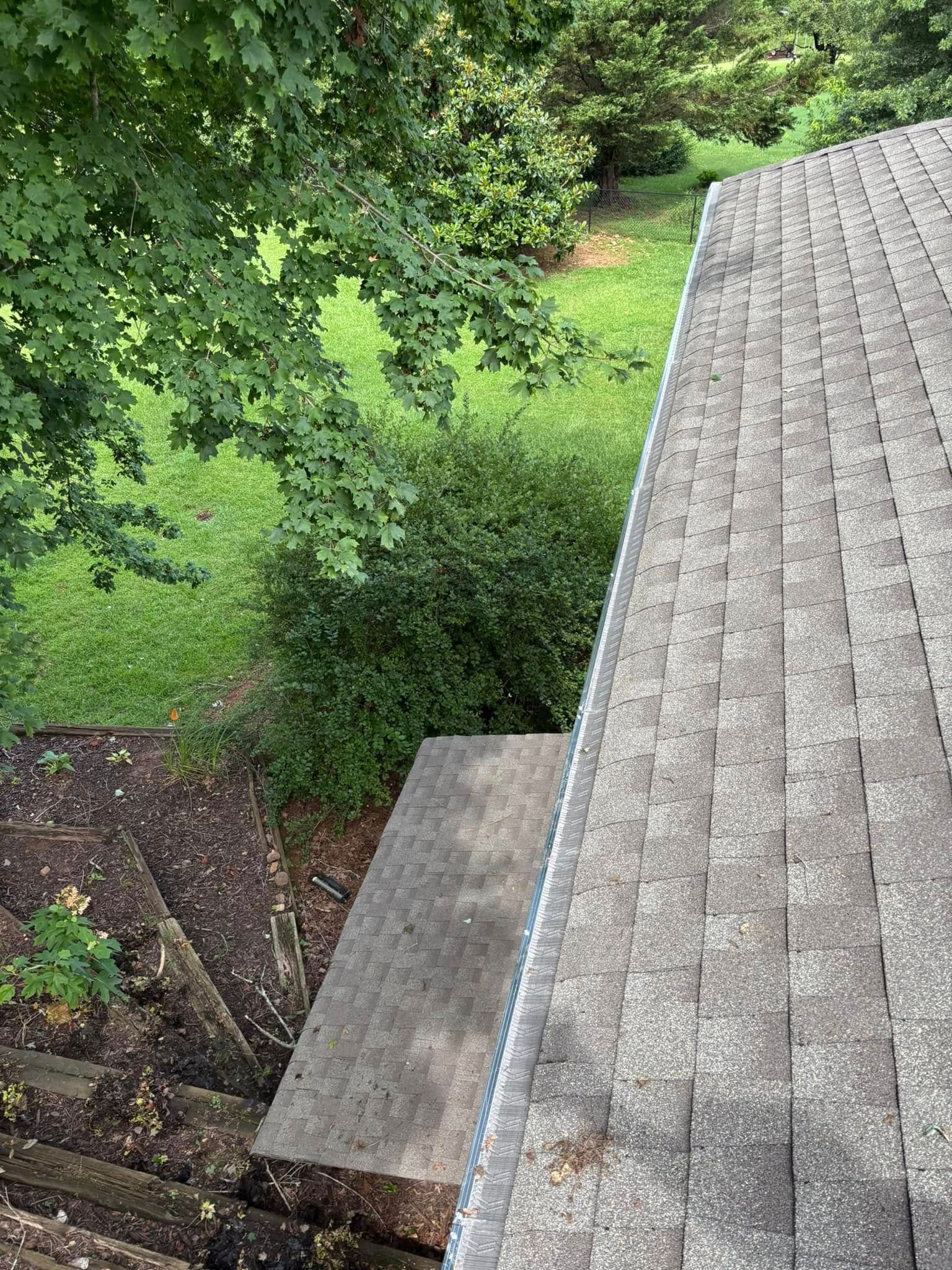 View from a roof, looking down at foliage, stairs, and part of another roof. Green grass and trees are in view.