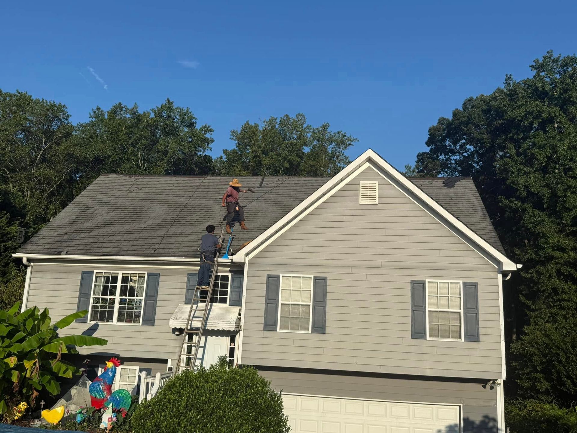 Two workers on a gray-roofed house, one on the roof, one on a ladder. Blue shutters, clear sky.