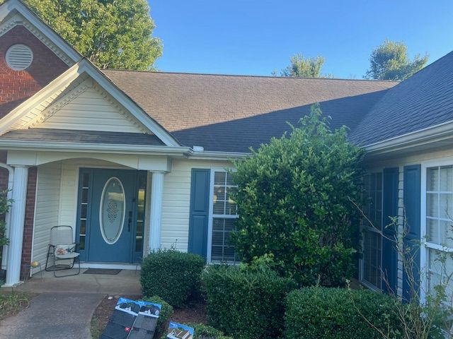 A light-yellow house with blue door, shutters, and dark roof. Green bushes and a blue sky are visible.