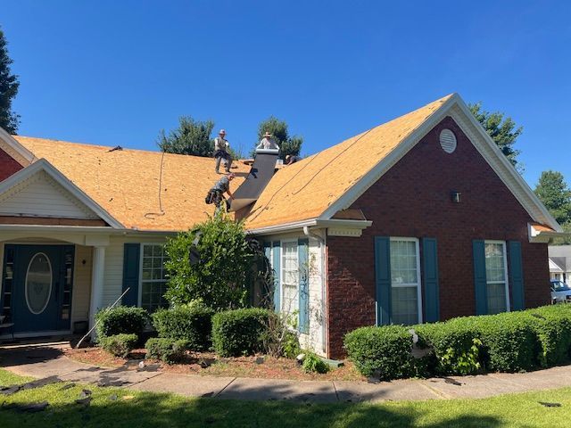 Roofers on a house with exposed sheathing; brick and light blue siding. Bright, sunny day.