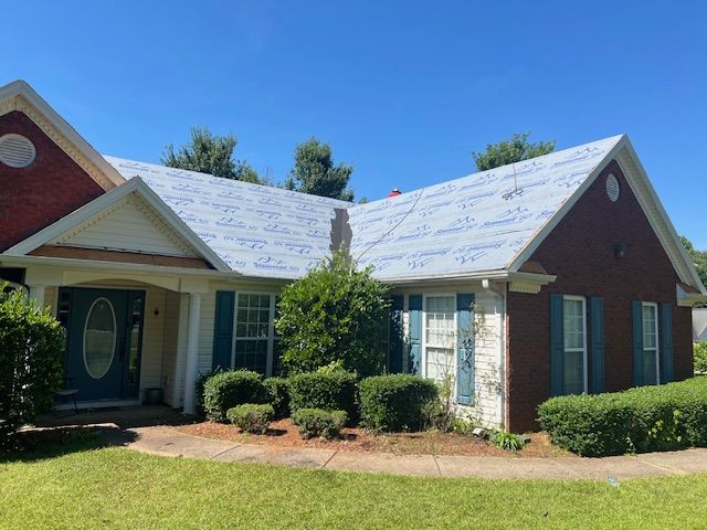 House with roof partially covered with blue protective sheeting. Brick exterior, blue shutters, and bushes.
