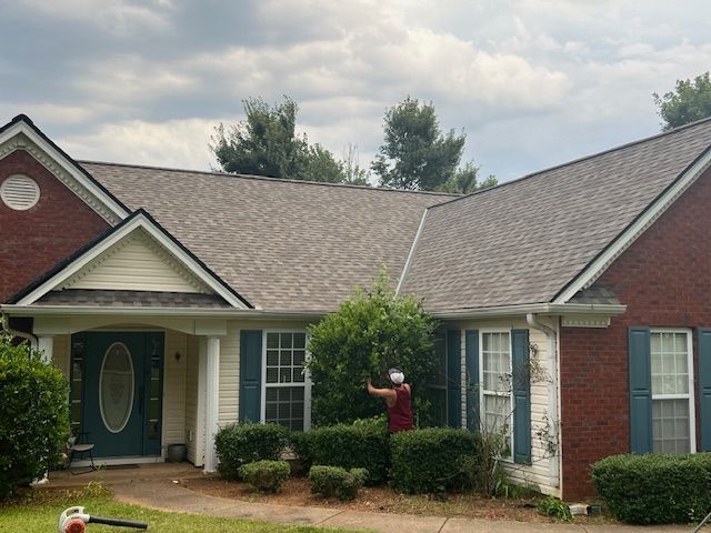 Person trimming hedges in front of a brick house with blue shutters under a cloudy sky.