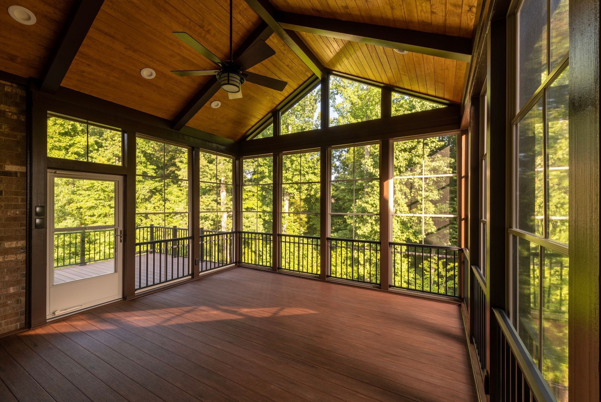 Screened porch with dark wood floor, exposed beams, and floor-to-ceiling windows overlooking a green forest.