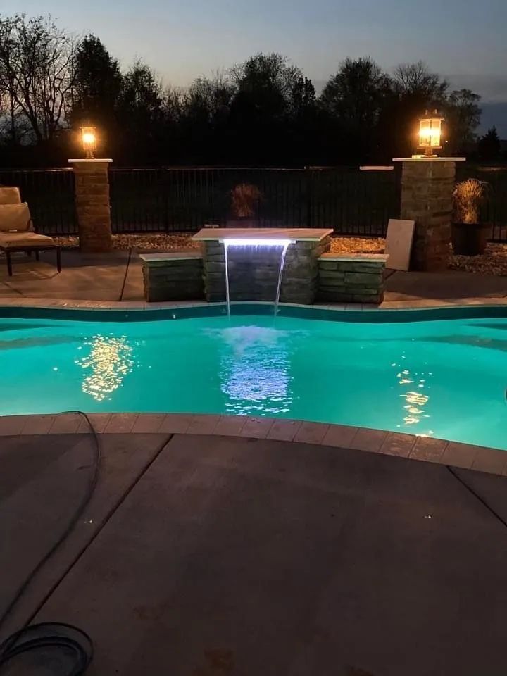 A backyard swimming pool at dusk with a stone water feature illuminated by lights, flanked by two lit stone pillars.