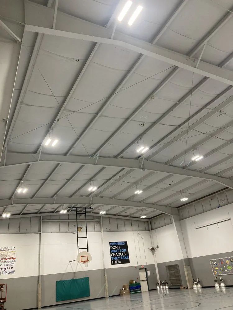Interior view of a gymnasium with a basketball hoop, white walls, and bright ceiling lights.